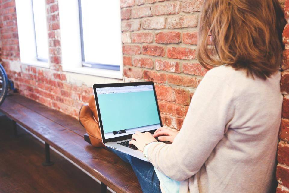 Vrouw werkt op een laptop in een vensterbank, met een bakstenen muur achter haar